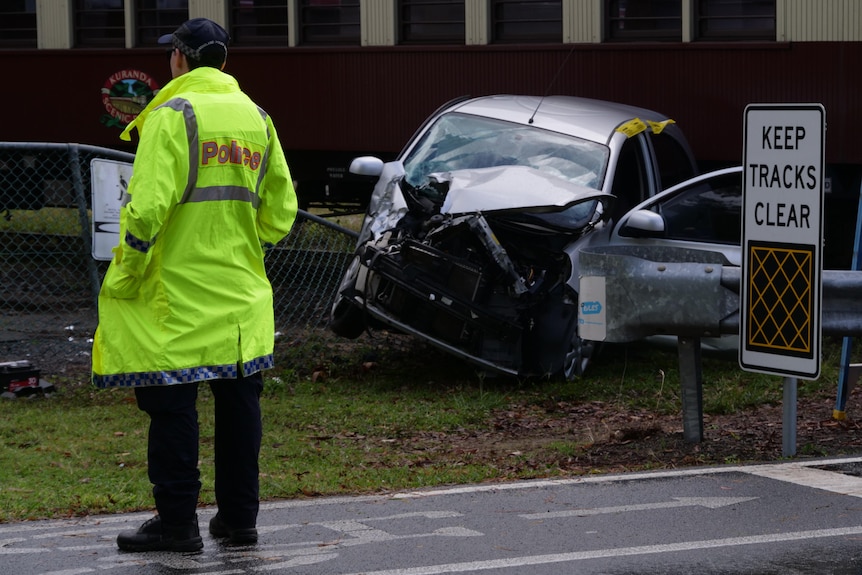 A police officer in a hi-vis jacket stands next to the wreckage of a silver hatchback on a railway track