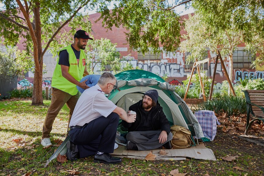 salvational army officer and vested volunteers give cup of coffee to man in a beard sitting in a tent in a parkh a 