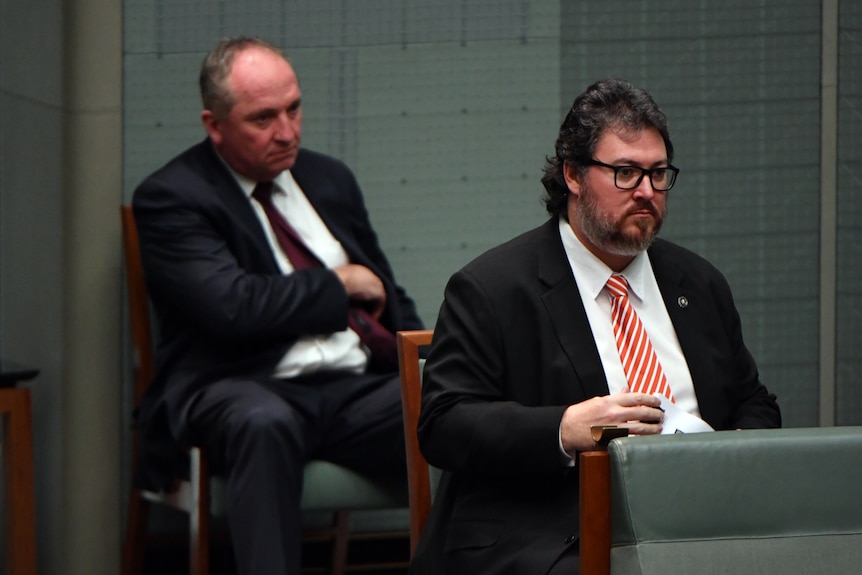 A man in a suit speaks in parliament, with another man in a suit behind him.