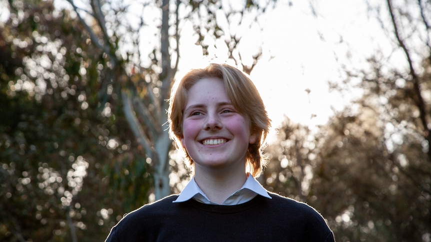 A young person with short blonde-brown hair stands outside, smiling.