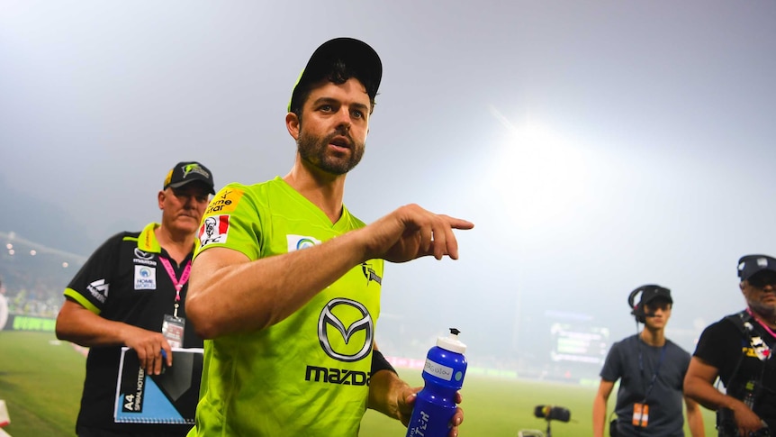 Callum Ferguson points as he holds a water bottle and trudges off as flood lights illuminate the smoke haze at Manuka Oval