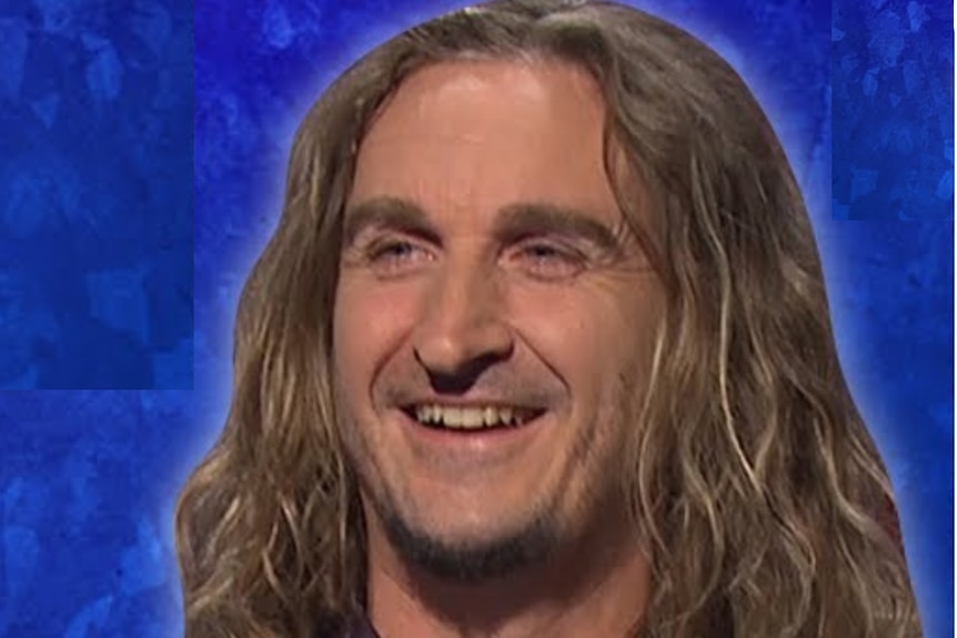 headshot of man with long hair smiling looking left of screen, with blue background