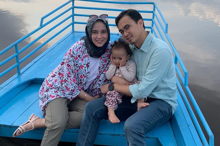 An Indonesian woman with a veil sits on a platform with her husband and child with a view of water in the background.