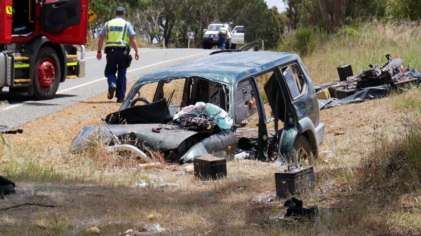 A crashed green 4WD sits in a ditch on the side of a road with its doors removed, with a fire truck and police officer behind.