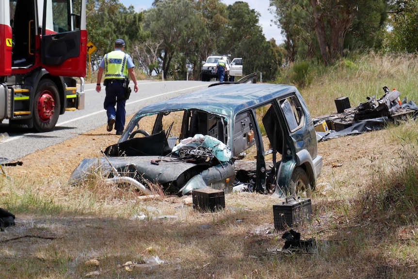 A crashed green 4WD sits in a ditch on the side of a road with its doors removed, with a fire truck and police officer behind.