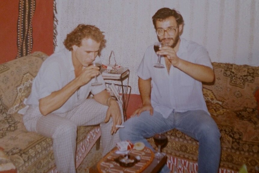 Two men drink wine as one makes a cheers gesture towards the camera.