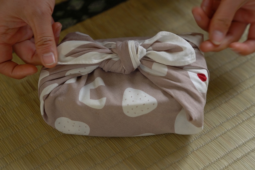 A small bento box in the shape of a rectangle is wrapped in light pink with dottie fabric. It' also has a bow tie knot on top. 