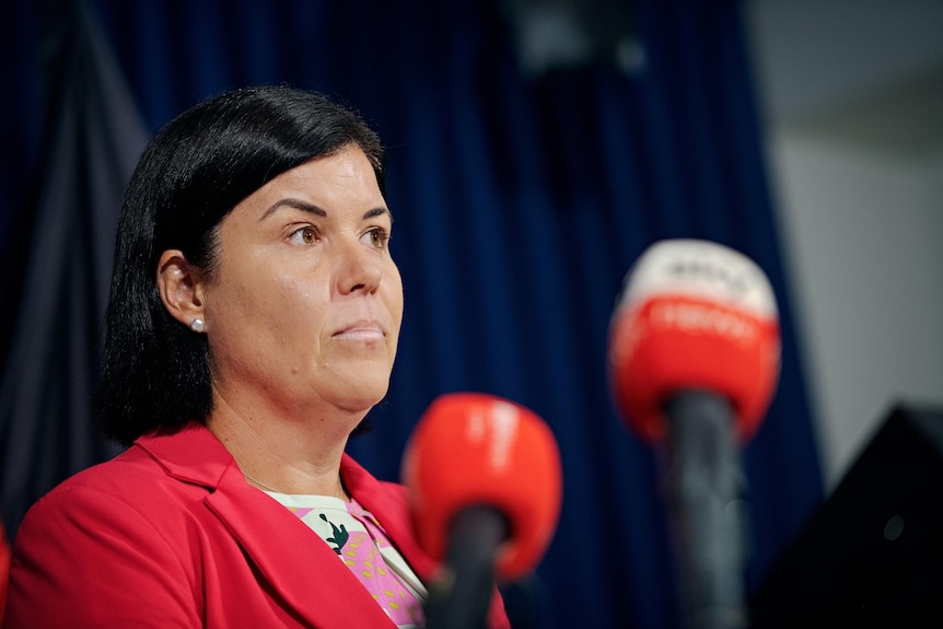 a woman with short dark hair wearing a pink blazer speaks at a lectern