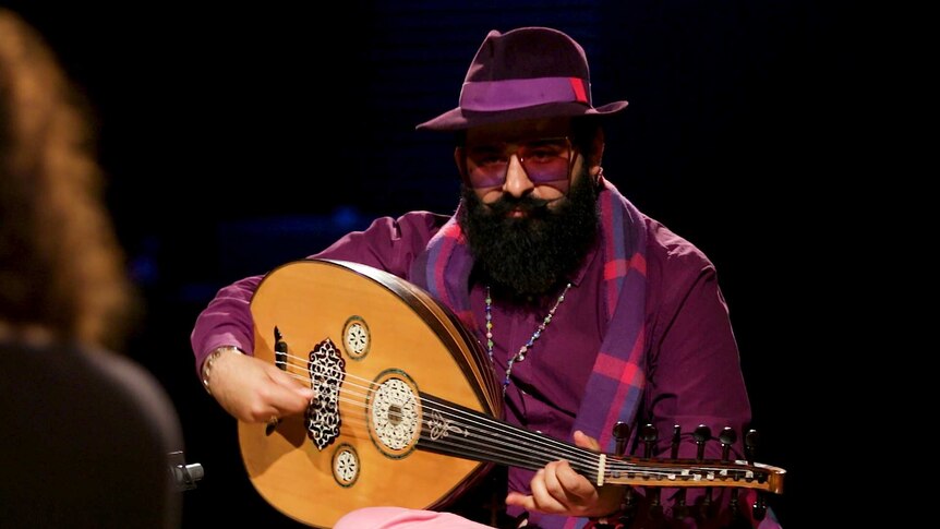 Oud player Joseph Tawadros plays while sitting on a chair. He wears a purple fedora, glasses, shirt and a matching plaid scarf.