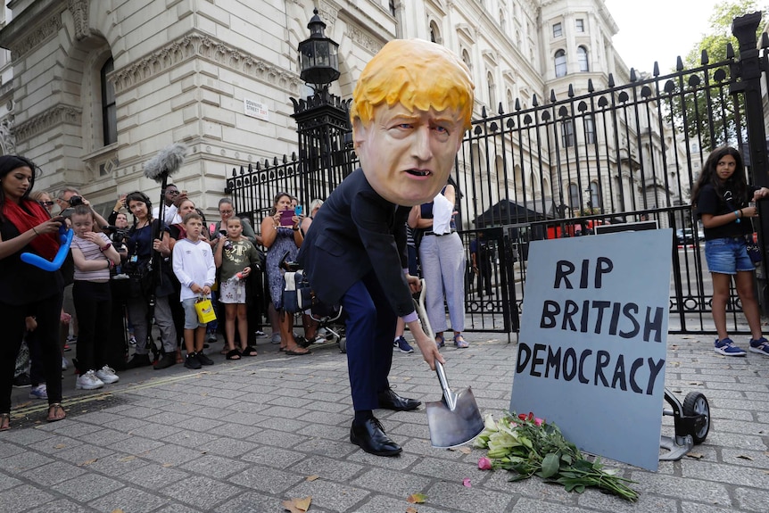 Man in large Boris Johnson head with a shovel and flowers and sign reading: 'RIP British Democracy'
