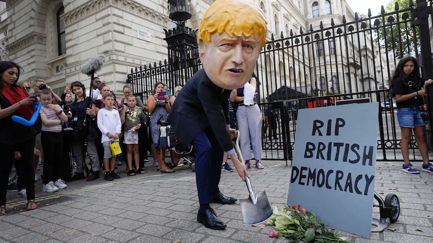 Man in large Boris Johnson head with a shovel and flowers and sign reading: 'RIP British Democracy'