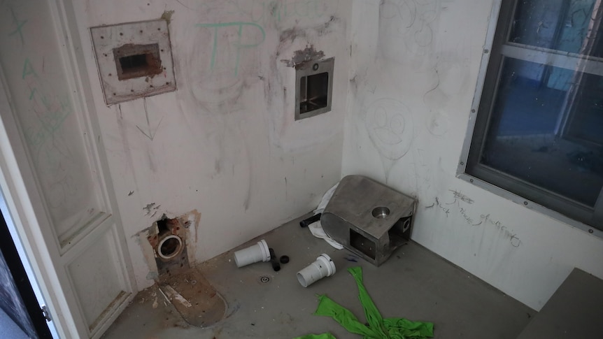 Graffiti and holes in the walls in a prison cells with items strewn on the floor