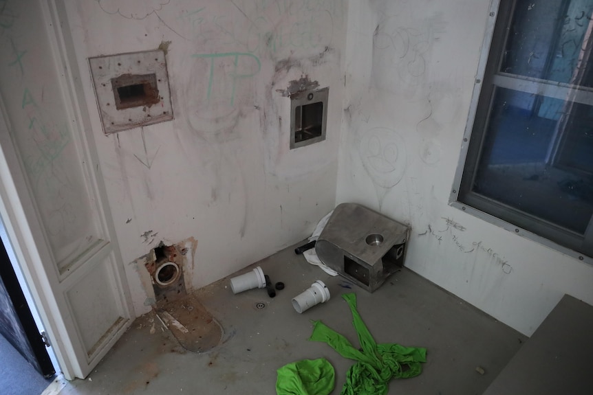 Graffiti and holes in the walls in a prison cells with items strewn on the floor