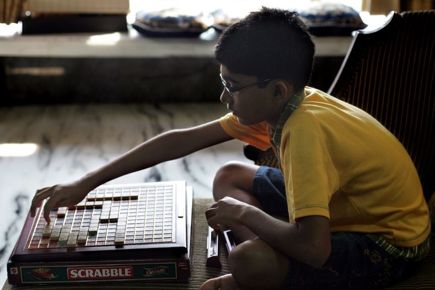 A boy in yellow shirt and glasses reaches over a Scrabble game board to move a small letter tile.
