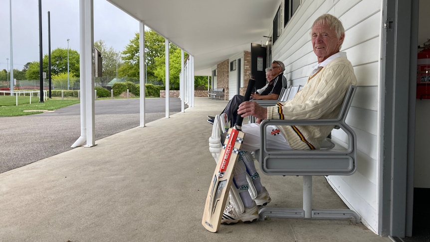 Older man sitting in his cricket whites with a bat in hand looking at the camera.