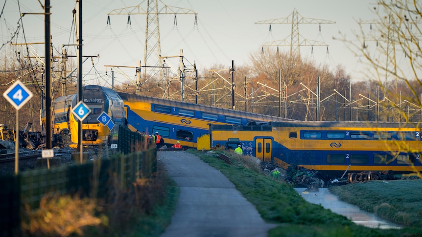 A cycle path is pictured with a yellow and blue derailed train on it, on the right side is a canal with another carriage in it.