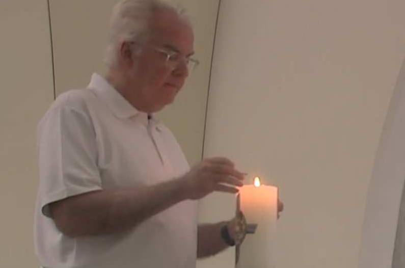 Older man with grey hair and glasses, wearing white shirt and black pants lights a candle