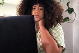 A mixed race woman sits down and looks at a laptop screen, seen from the back. Her short curly hair is out, she wears check top 