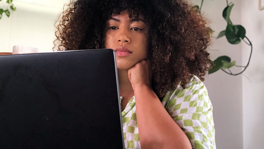 A mixed race woman sits down and looks at a laptop screen, seen from the back. Her short curly hair is out, she wears check top 