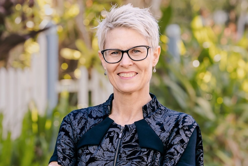 A woman with short, grey hair, wearing glasses, stands smiling with her arms crossed.