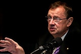 Harold Mitchell, wearing glasses and suit with brown hair, looks past a microphone, gesturing with his right hand.