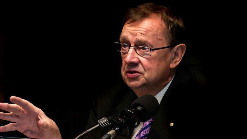 Harold Mitchell, wearing glasses and suit with brown hair, looks past a microphone, gesturing with his right hand.