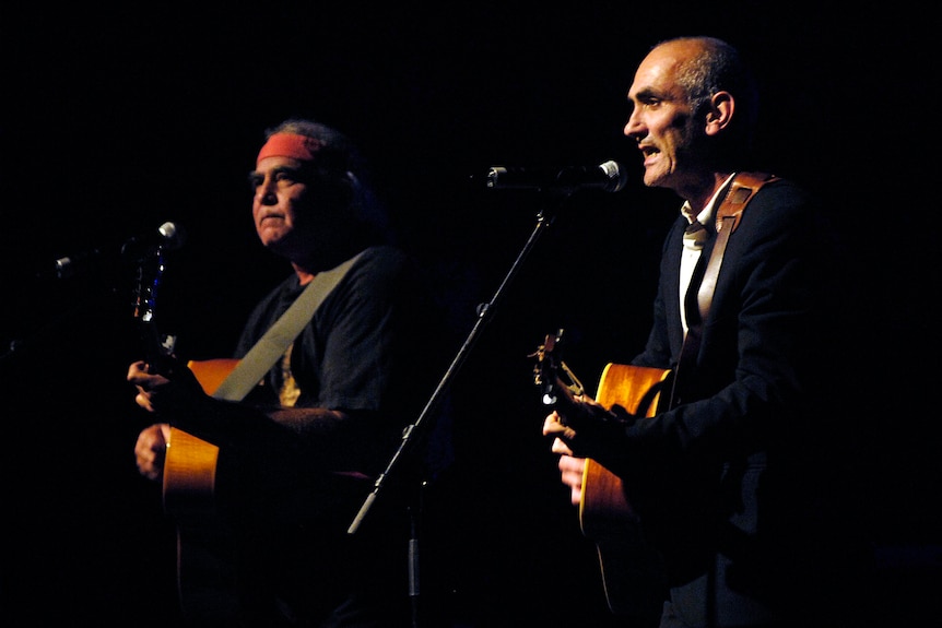 Two men sit together playing guitar and singing into microphones
