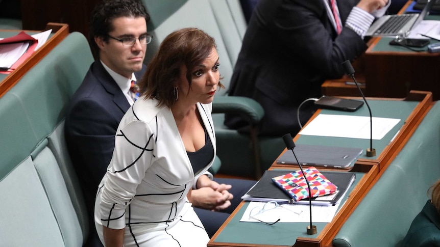 Anne Aly, hovering over her seat, has her eyes wide open and eyebrows raised. She is wearing a white and black dress.