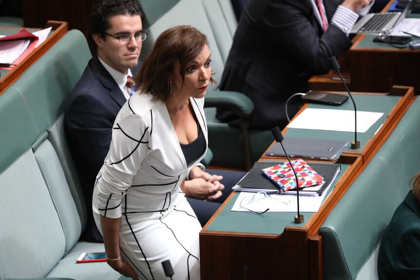 Anne Aly, hovering over her seat, has her eyes wide open and eyebrows raised. She is wearing a white and black dress.