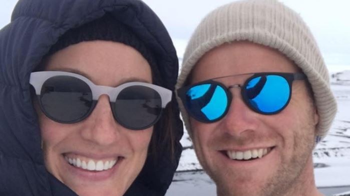 Amber Griffiths and Andrew Powell pose for a photo in beanies and sunglasses against a snowy landscape backdrop.