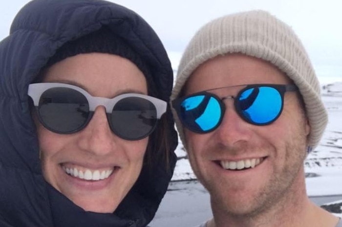 Amber Griffiths and Andrew Powell pose for a photo in beanies and sunglasses against a snowy landscape backdrop.