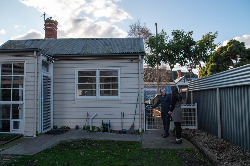 Two people stand outside, next to a fence that connects to the side of a weatherboard house