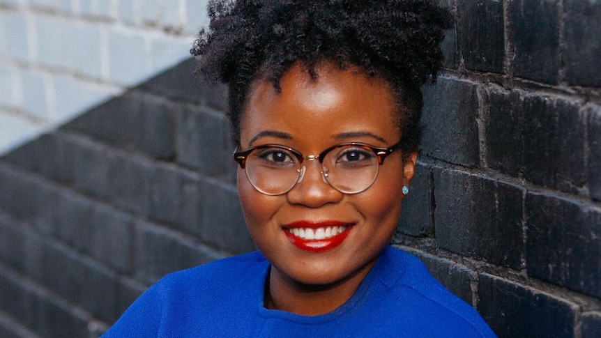 A woman with black curly hair and a bright blue top standing in front of a brick wall