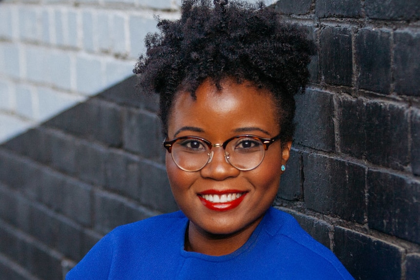 A woman with black curly hair and a bright blue top standing in front of a brick wall