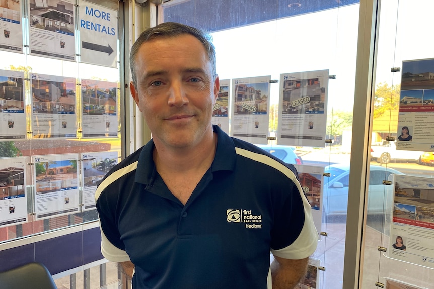 A man with short brown hair wearing a blue shirt stands in front of his office window, which is covered in ads for homes.