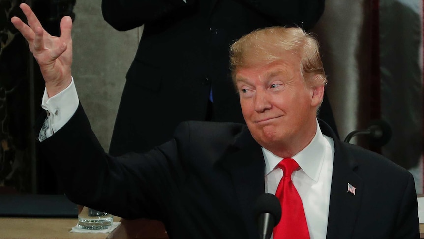 Donald Trump, wearing a suit, holds up his right hand and looks in that direction.