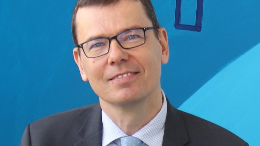 Man in suit and glasses smiles in front of blue background.