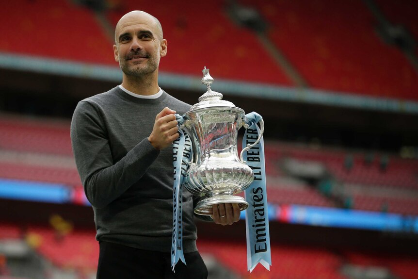 Pep Guardiola stands in an empty stadium holding the FA Cup