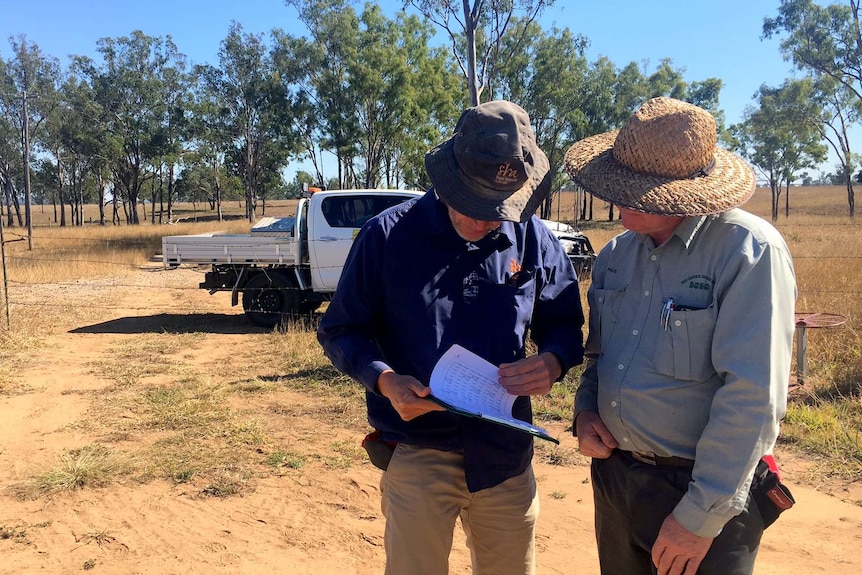 Two men with their heads down look at a clipboard in a field