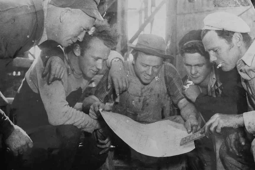 Old monochrome image of five crouching workmen huddle around a building plan.