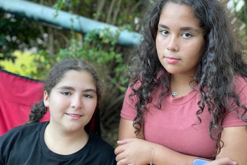 Two sisters pose for a photo outdoors. The older of the girls has her arms crossed and the younger is smiling.