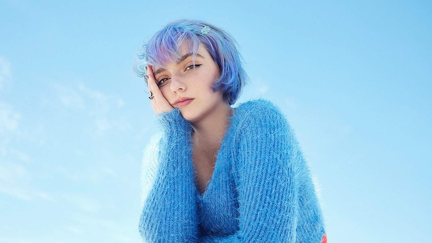 Photo of singer merci, mercy sitting with her hand on her face with blue hair and fluffy jumper