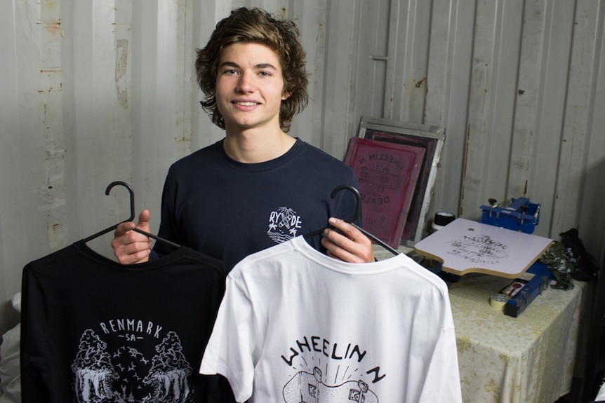 A teen boy with brown hair stands in a shipping container holding two t-shirts.