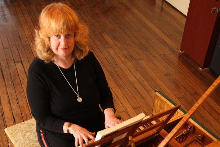 A woman looks up at the camera. She is seated at a harpsichord with her fingers on the keys.
