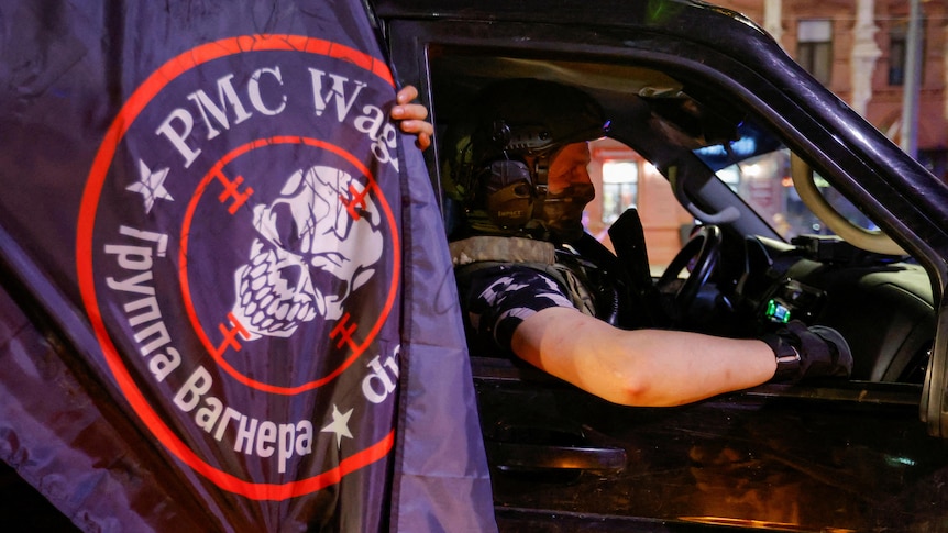 A black flag emblazoned with a white skull and WAGNER PMC hanges next to a 4WD. The driver wears camouflage face mask