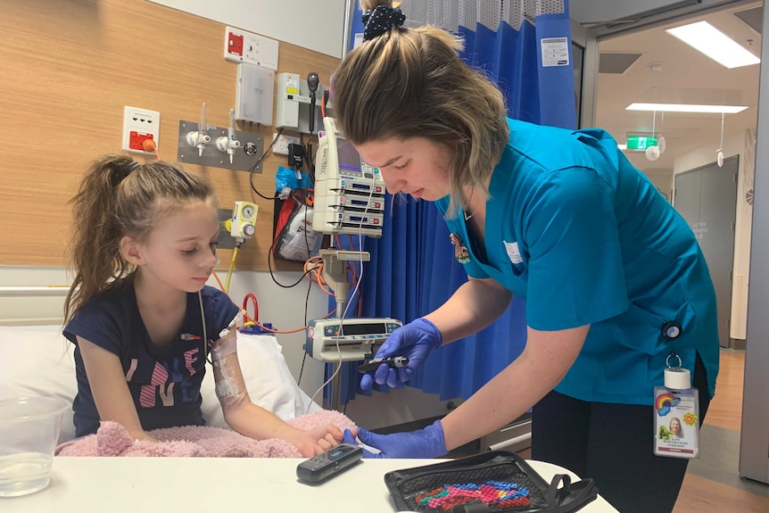 Hayley Minson-Rivers sitting in a hospital bed with a nurse standing next to her