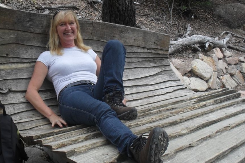A woman with blonde hair wearing a white shirt sits on wood. There is a tree and foliage in the background.