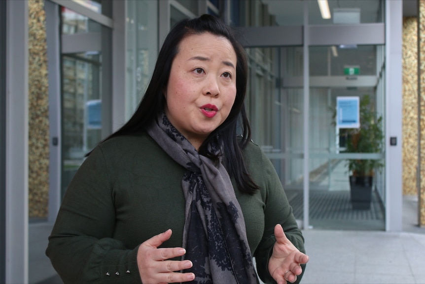 An Asian woman wearing a scarf, stands with her hands out, gesturing as she speaks.