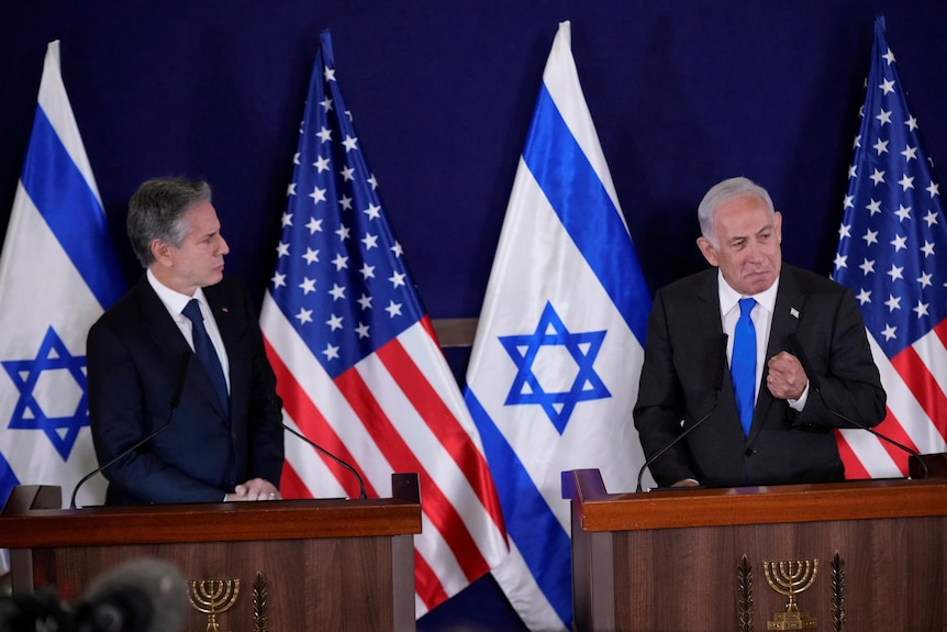 Two men in suits, behind podiums, with Israel and US flags in the background, speak at a press conference