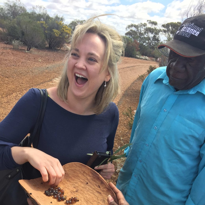 Indigenous elder holds a calabash of honey ants which a laughing woman from conference industry takes one and laughs